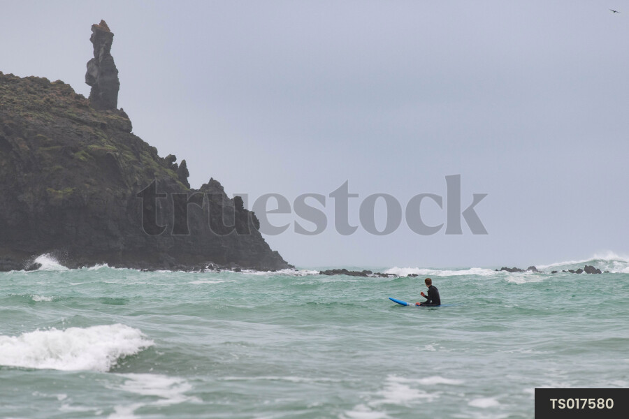 Man sitting on surfboard by rocky coastline