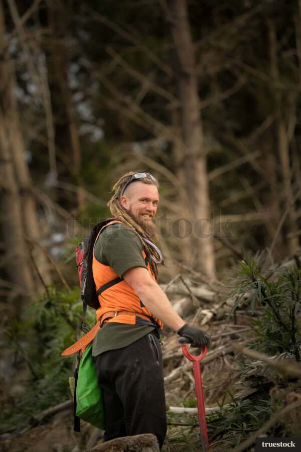 Male forester planting new trees wearing reflective clothing carrying bag full of trees and shovel