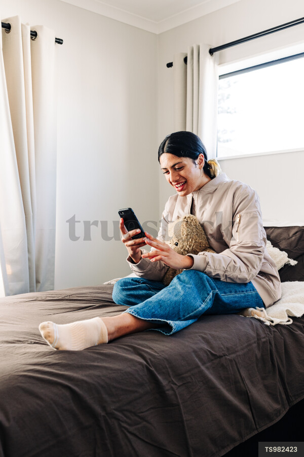 Young woman with smartphone and teddy bear sitting on bed